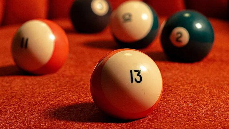 Darstellung der Zahl 13 durch eine Billiardkugel, die mit einer 13 beschriftet auf einem roten Sofa liegt. Im Hintergrund liegen weitere Billiardkugeln, deren Zahlen aber nicht erkennbar sind.