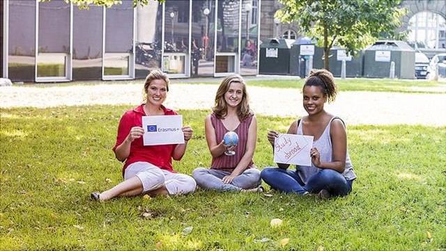 Drei Studierende sitzen auf der Wiese und halten Schilder mit der Aufschrift "Erasmus" und "Study Abroad" sowie einen Globus