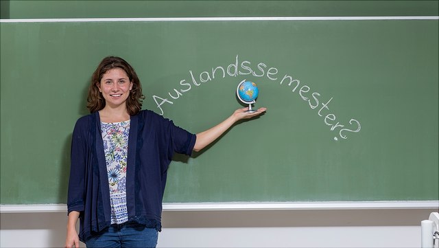 Studieren im Ausland Person steht vor einer grünen Kreidetafel auf der "Auslandssemester?" geschrieben steht. Die Person hält einen kleinen Globus in der Hand