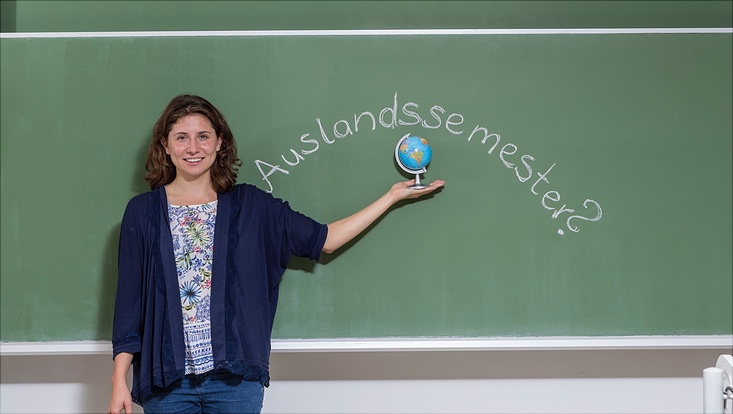 Eine junge Frau steht vor einer Tafel mit dem Anschrieb "Auslandssemester?" und hält einen Globus in ihrer ausgestreckten Hand.