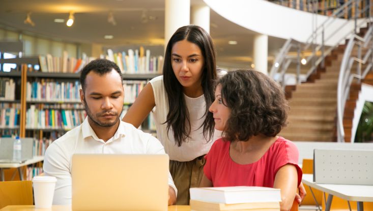 Drei Studierende in Bibliothek schauen auf Laptop