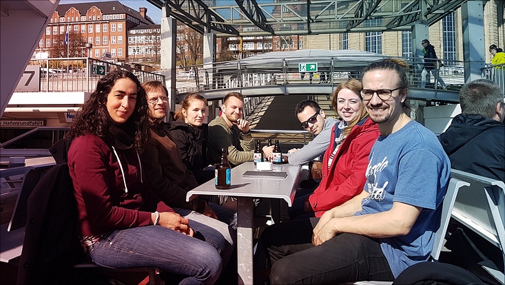 Eine Gruppe von 7 Studierenden sitzen um einen Tisch auf einer Fähre herum und schauen Richtung Kamera.