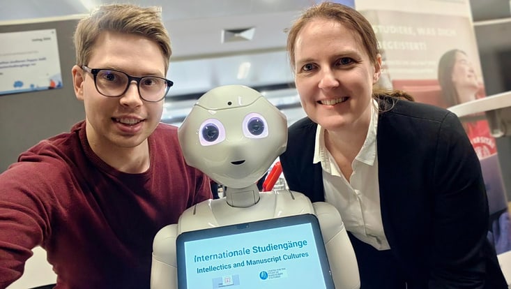 Zwei Personen stehen neben einem humanoiden Roboter mit Tablet an einem Informationsstand zu internationalen Studiengängen. Auf dem Bildschirm steht „Internationale Studiengänge – Intellectics and Manuscript Cultures“.