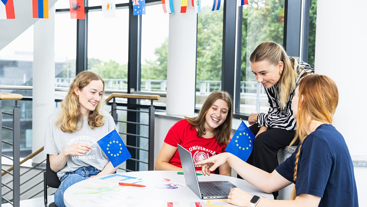 Drei Studentinnen sitzen zusammen am Tisch, lächeln und halten Europa-Fahnen, eine Studentin zeigt einer weiteren etwas am Laptop