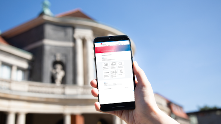 Hand hält ein Smartphone mit verschwommener Ansicht der DL-Büro Website. Im Hintergrund verschwommen das Hauptgebäude der Universität