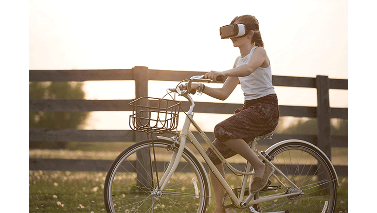 Ein Mädchen fährt an einem sommerlichen Tag auf einem Feldweg Fahrrad. Sie trägt eine Virtual Reality Brille.
