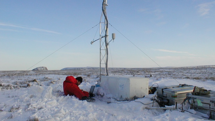 Ein Forscher in einem roten Schneeanzug sitzt im Schnee. Neben ihm eine Messstation. Symbolisch für Arktis, Permafrost und Meereis.