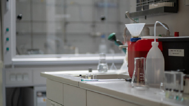 Labortisch mit rotem Kanister und verschiedenen Gläsern im Vordergrund und Chemikalienabzug im Hintergrund.