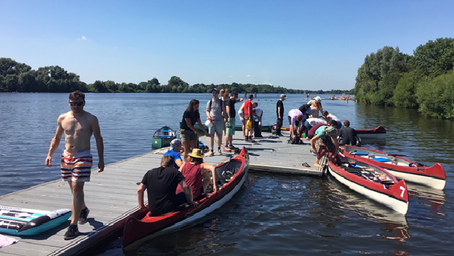 Holzsteg im Wasser, an dem drei rot-weiße Kanus angelegt haben und auf dem eine Gruppe von Menschen steht und sitzt.