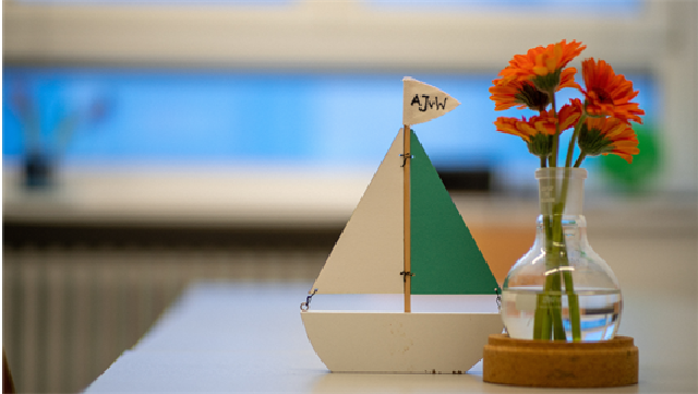 Weiß-türkises Holzsegelschiff und Glasvase mit vier roten Blumen auf einem unscharf zu sehenden Tisch.