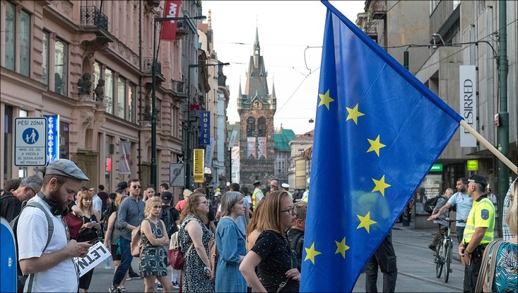Menschen stehen versammelt auf einem Platz. Im Vordergrund ist eine EU-Flagge zu erkennen.