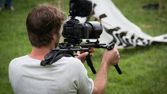Ein Mann von hinten. Auf seiner linken SChulter eine Kamera. Vor ihm ein Zelt.
