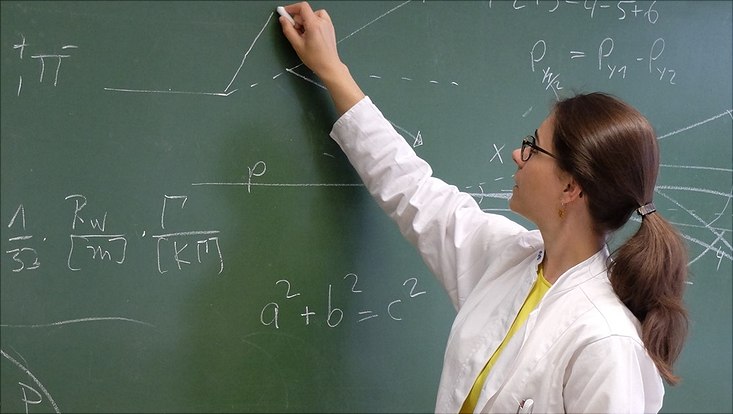 A young woman in a white coat writes formulas on the blackboard