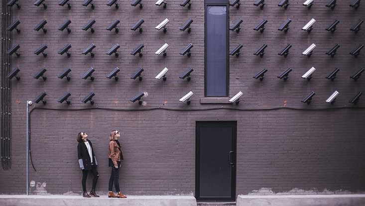 Zwei Frauen stehen vor einer Backstein-Hauswand und schauen auf zahlreiche Überwachungskameras, die dort monitert sind