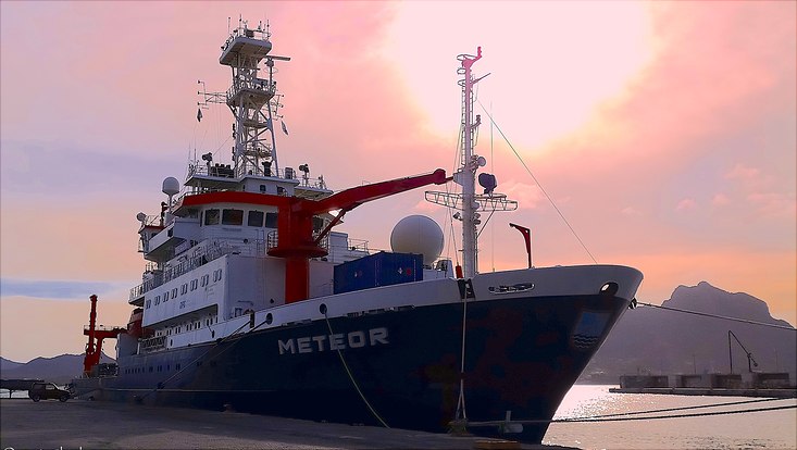 Das Forschungsschiff in einem Hafen. Im Hintergund ist ein rot schimmernder Sonnenuntergang zu sehen.
