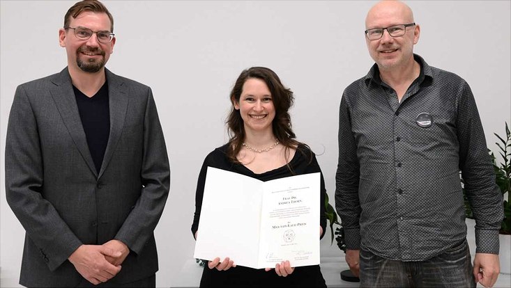 A woman and two men stand smilingly in front of a white wall. The woman hold a certicicate. hält eine Urkunde.