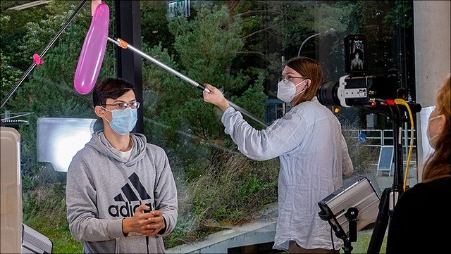 A woman holds a pink ballon on a stick above a boy's head, who is looking into a camera, held by a girl on the right.
