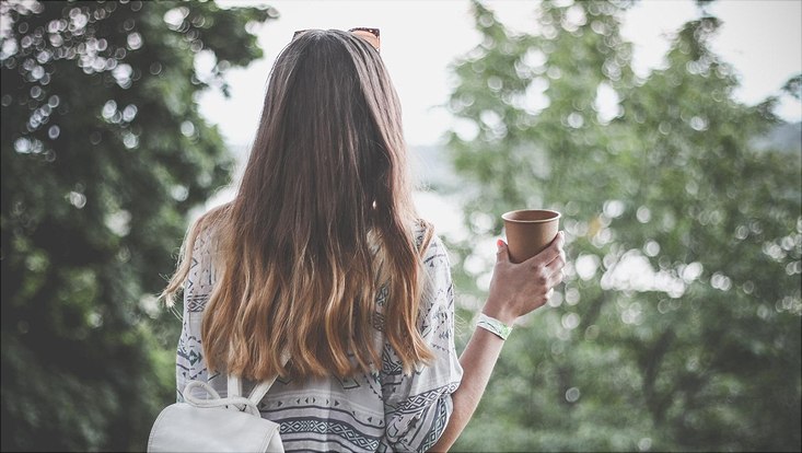 Frau, von hinten abgelichtet, mit Kaffeebecher in der Hand, draußen