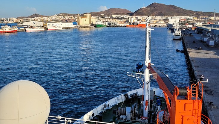 Zu sehen ist das Schiff, wie es im Hafen von Las Palmas liegt. Im Hintergrund sind Berge zu sehen.