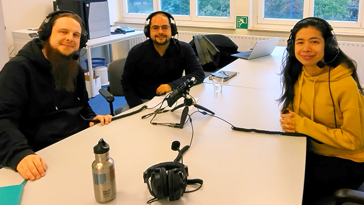 Carlos Tello and Alejandra Gómez Correa sitzen mit Podcast Coach Michael Büker an einem Tisch und tragen Kopfhörer. In der Mitte des Tisches steht ein Mirkofon.