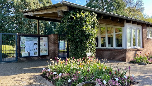 Besuchereingang des Botanischen Gartens. Im Vordergrund ein mit bunten Blumen bepflanztes Beet, im Hintergrund ein Backsteingebäude mit weißer Fensterfront.