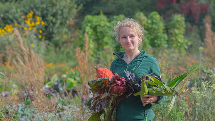 Bo bei Ernte Eine junge Gärtnerin steht inmitten einer üppig blühenden Staudenwiese und hat dieverse Gemüse auf dem Arm, die sie gerade geerntet hat