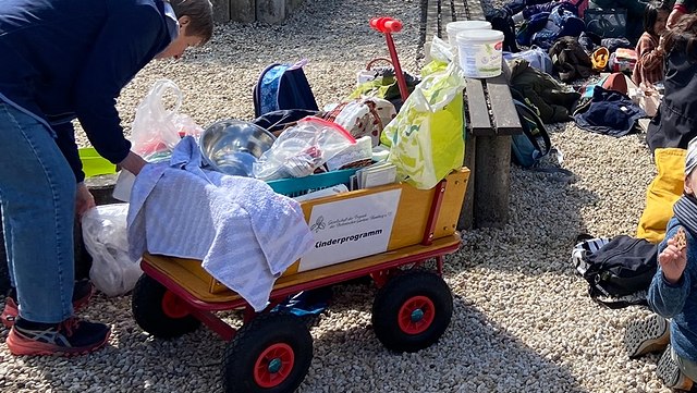 teaser kinderprogramm 640x361 Ein mit allerlei Bastelkram beladener Bollerwagen im Vordergrund, im Hintergrund eine Gruppe Kinder