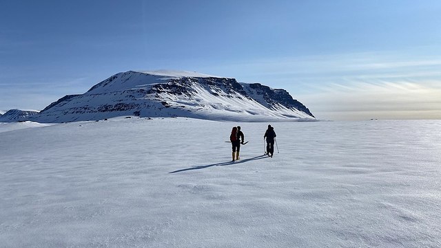 Zwei Menschen laufen mit Skieern über eine schneebedeckte Fläche. Im Hintergrund sieht man einen Berg aufragen.