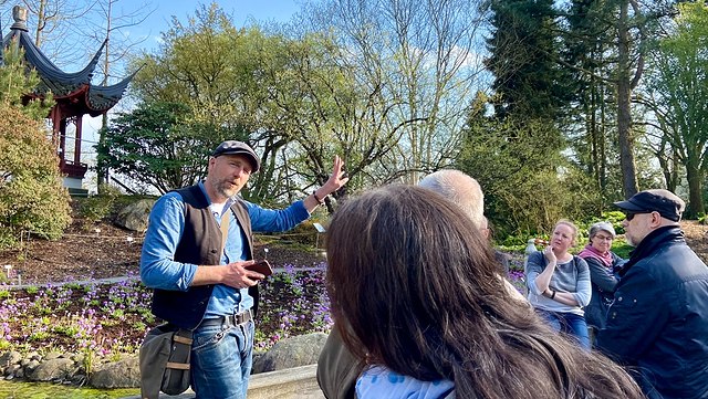 individuelle gruppenführungen-640x361 Frontansicht eines jungen Mannes der gerade etwas erklärt, Rückeansicht einiger Zuhörer:innen, das Ganze im Botanischen Garten Hamburg