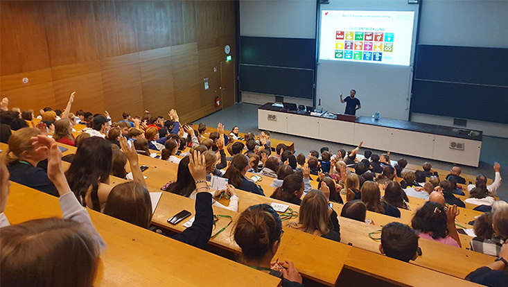 Auf dem Bild sind Schüler*innen von hinten in einem Vorlesungsaal zu sehen. Einige von ihnen heben ihre Hand und melden sich. Vorne steht Dr. Leonard Borchard und erklärt den Schüler*innen etwas über die Nachhaltigkeitsziele.
