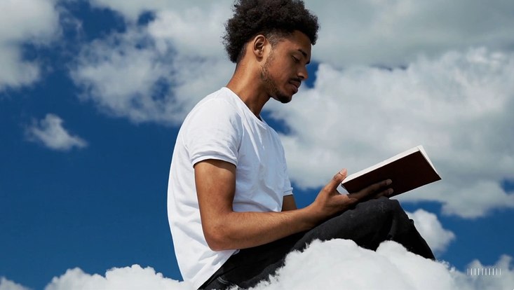 A young man at his 20s is sitting on a piece of cloud in the sky, reading a book.