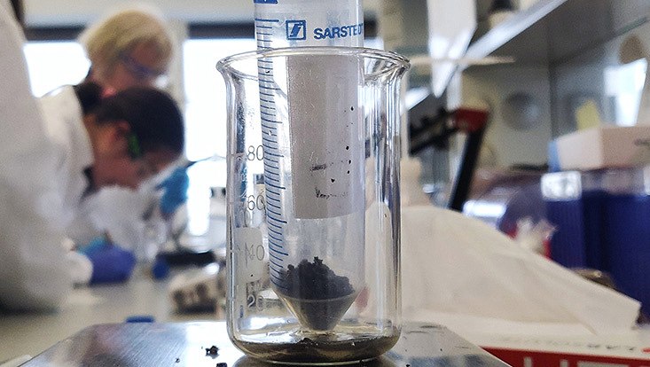 Zu sehen ist ein Versuchsaufbau mit einem Becherglas in dem eine Spritze mit Erde drin ist. Im Hintergrund sind Schüler:innen zu sehen.
