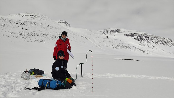 two scientists taking gas samples from different snow depth via a tube inserted to the snow. Snow-covered landscape with hills in the back.
