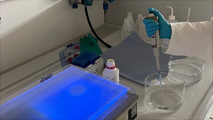 img-6929-733x414 Auf der rechten Bildseite sieht man eine Person beim Pipettieren einer farblosen Flüssigkeit. Auf der linken Bildseite steht ein flaches Gerät das blau aufleuchtet.