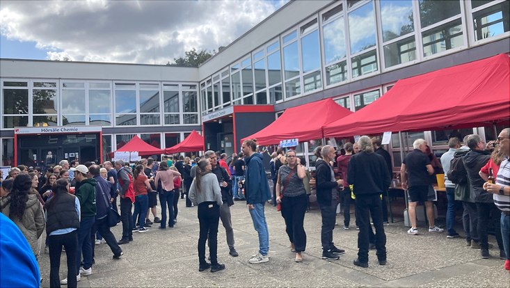 img-6869-733x414 Im Hintergrund erkennt man ein Gebäude. Davor sind verschiedene Pavillions zu sehen und viele Menschen, die sich unterhalten.