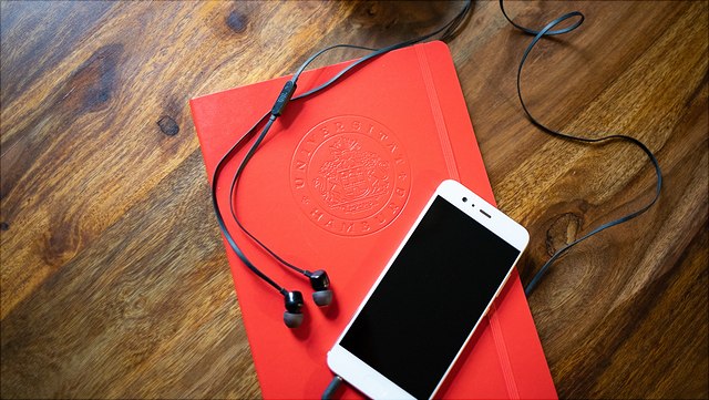 201116-symbolbild-handydisplay-6600-640x361 Ein weißes Smartphone mit dunklem Bildschirm liegt auf einer roten Uni Hamburg mappe. Es sind schwarze Kopfhörer an das Smartphone angeschlossen, alles liegt auf einem dunklen Holztisch.