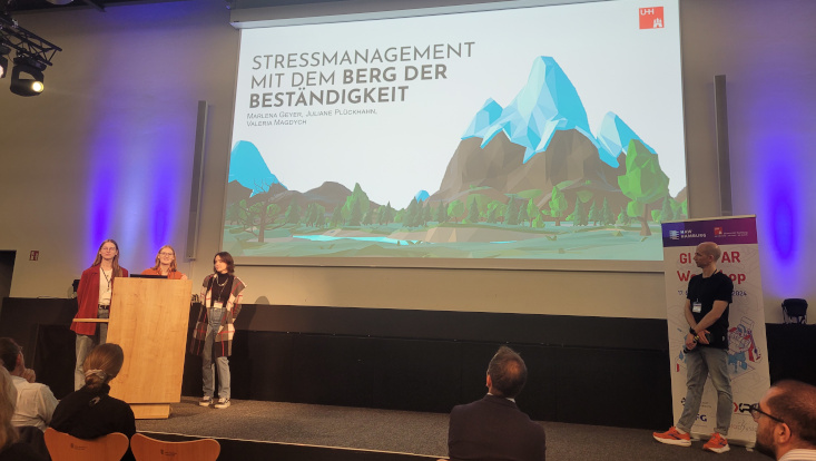Three people are presenting their work on a stage, with the presentation featuring a large mountain.