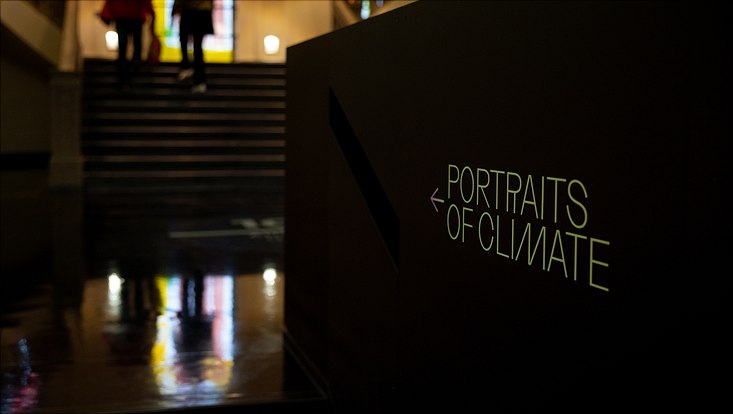 bild1-3770-portraits-of-climate Das Bild zeigt den Eingang zur Ausstellung. Im Hintergrund sieht man Treppenstufen, im Vordergrund rechts steht an einer Wand "Portraits of  Climate" mit einem Pfeil richtung Treppen. Die Wand ist dunkel und die Schrift ist goldfarben.