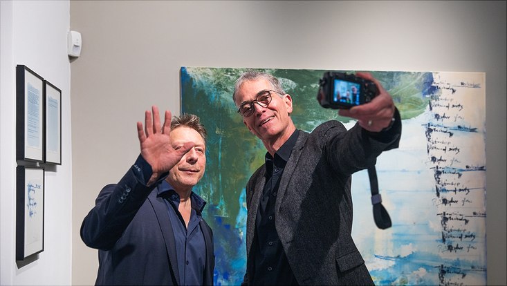 bild3 Zwei ältere Männer machen ein Selfie mit einer Kamera. Sie lächeln und der, der nicht die Kamera hält, winkt. Beide tragen Anzüge.