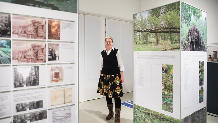 bild5-3956 Eine Frau mit buntem Rock steht zwischen zwei Ausstellungssäulen auf denen Fotos von Bäumen abgebildet sind.