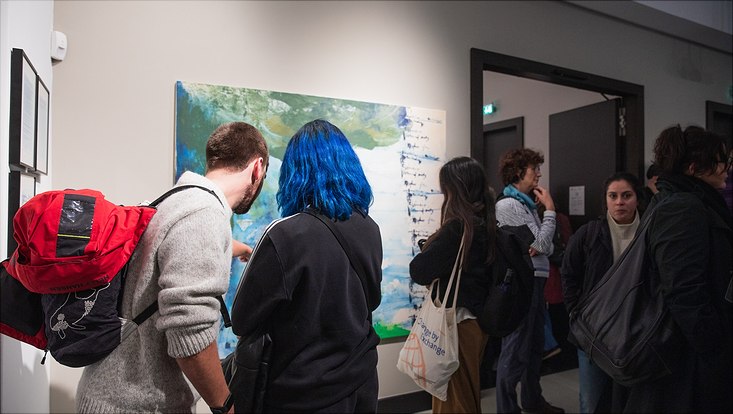 bild12 Man sieht mehrere Ausstellungsgäste, die sich die Kunstwerke angucken. Im Fokus sind eine Person mit blauen Haaren und eine mit kurzen brauen Haaren, die sich ein abstraktes Gemälde angucken, welches grüne und blaue Farbverläufe hat
