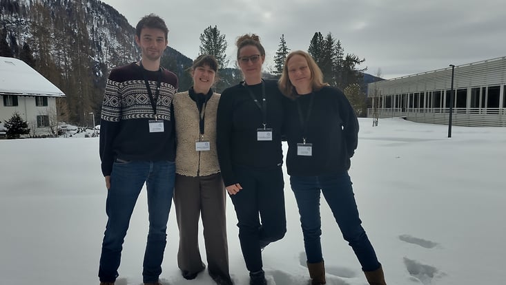 four persons standing on snowy ground with a mountain in the background and a large building on the right edge of the picture
