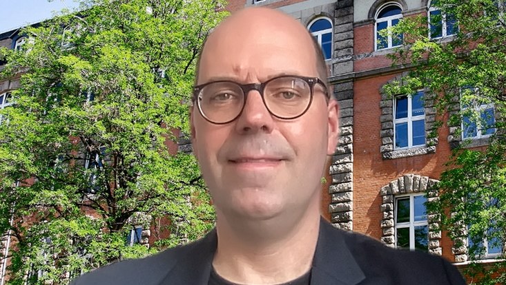 man with glasses in front of trees and brick building