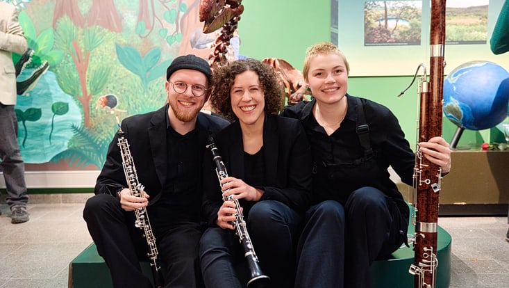 2025-02-cen-graduiertenfeier--8864-733x414 Die Mitglieder:innen des Trio d'Anches heureux sitzen ihren Instrumenten unter der Nachbildung eines Skeletts eines großen Laufvogels und lächeln in die Kamera.
