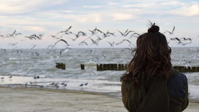 A person with long wavy hair and a top bun is standing at the beach. The sky is cloudy and there are many segulls flying over the water.