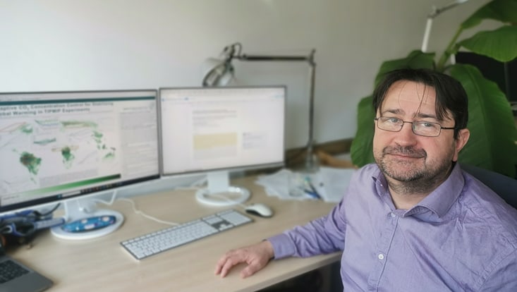 Scientist facing the camera and sitting next to the desk with two screens on it.