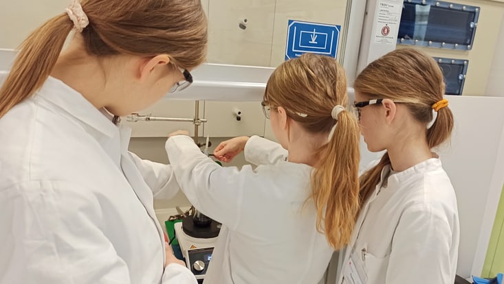 img-20250313-103135-733x414 Man schaut drei Schülerinnen im Chemielabor über die Schulter, die beim Experimentieren sind.