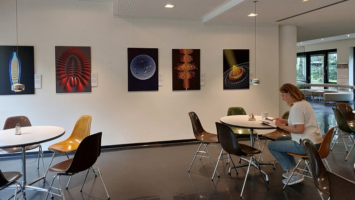 Blick auf fünf Bilder in der Cafeteria. Im Vordergrund sitzt eine Frau und liest.