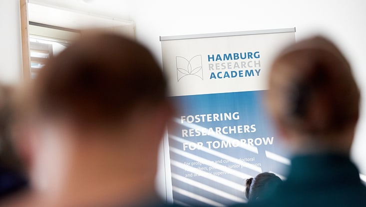 persons standing in front of a poster display of the Hamburg Reseach Academy
