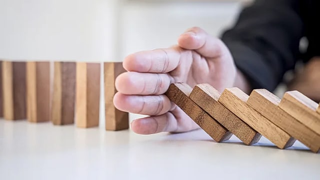 Auf dem Bild sind Holzdominiosteine zu sehen, die gerade dabei sind umzufallen und von einer Hand aufgehalten werden sollen.